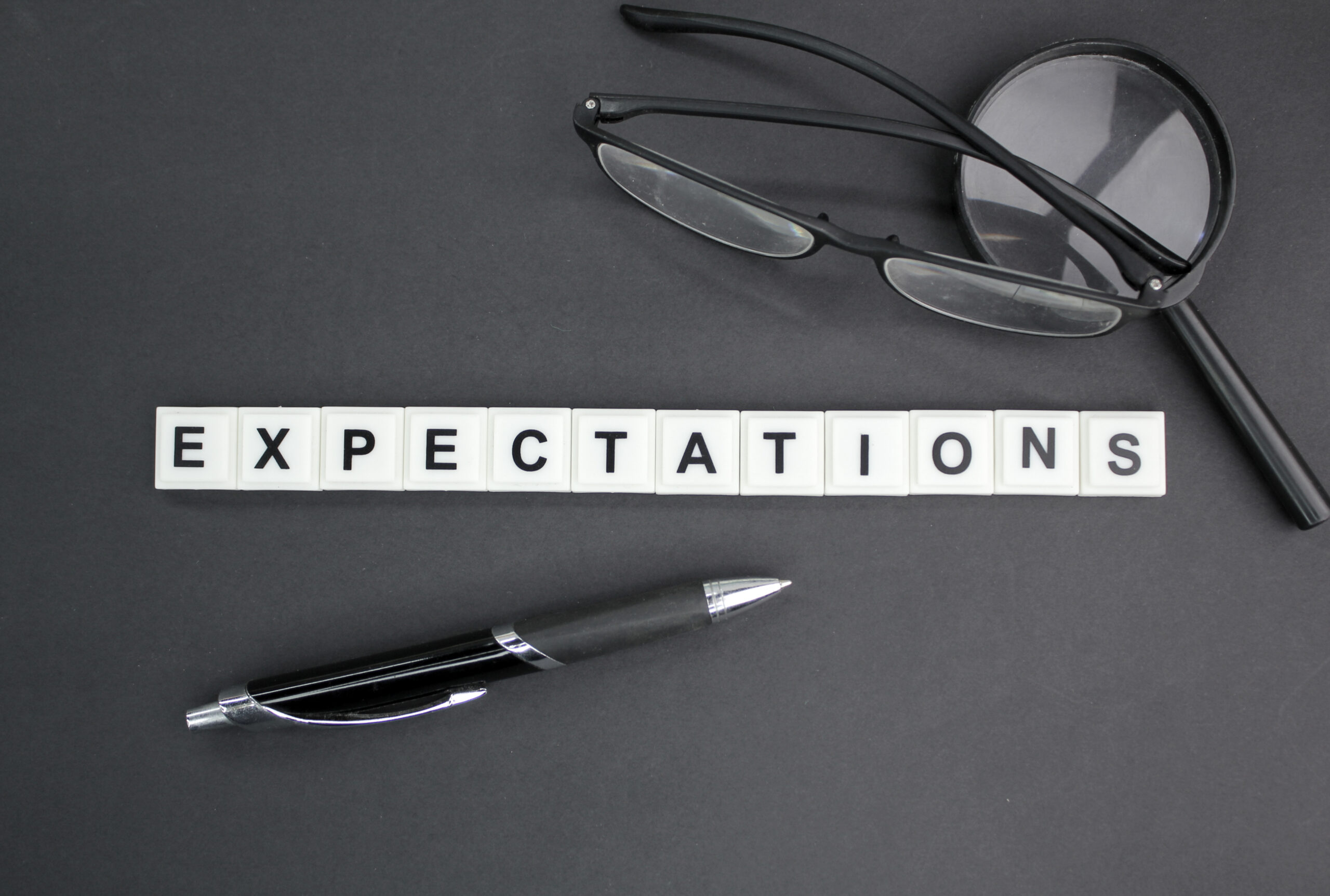 The word “EXPECTATIONS” spelled out on a dark background with glasses and office tools.